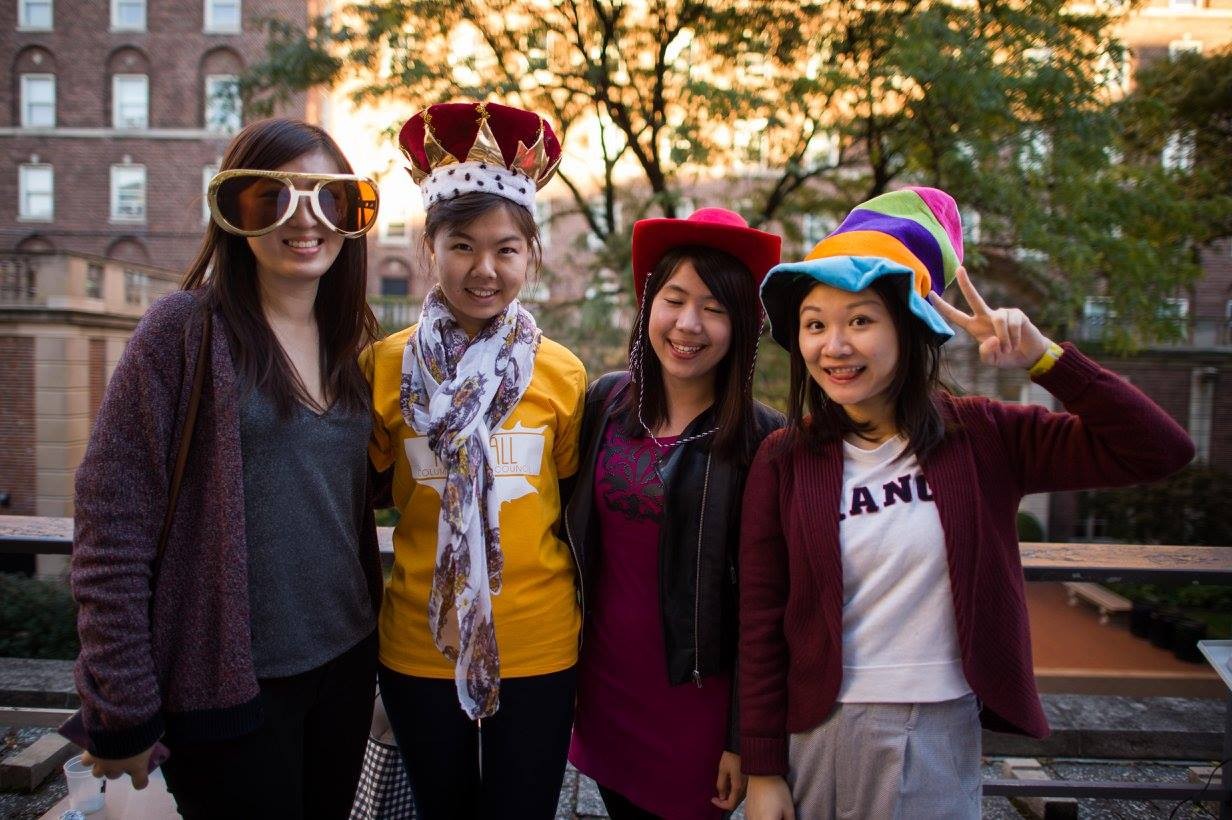 4 students posing for the camera with large glasses and silly hats such as a king's crown, cowboy hat, and mutli-colored floppy wizard hat.