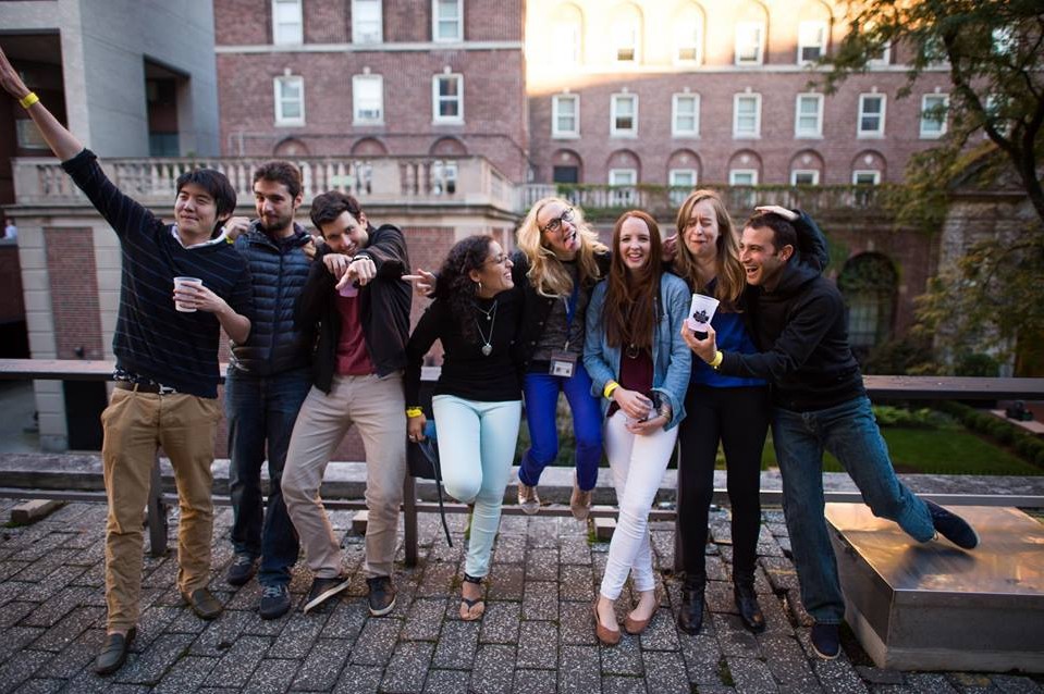 8 students posing for the camera with funny faces. One student is showing off their cup while another student is pointing to the camera. A student on the left is pointing off the image with a cup in his other hand.