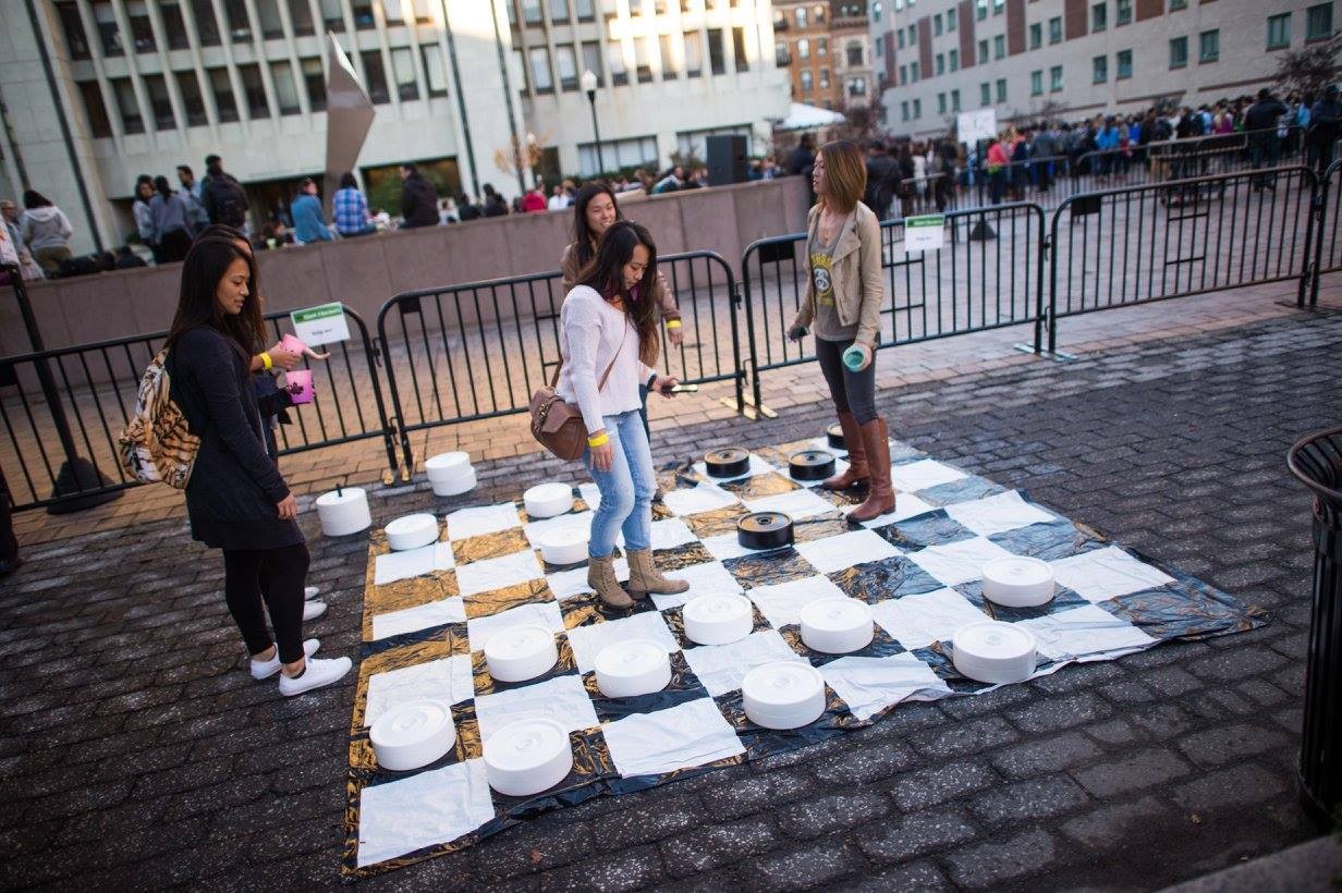 4 students playing Giant Checkers-- a black and white mat on the floor with students standing near large black and white wooden circles.